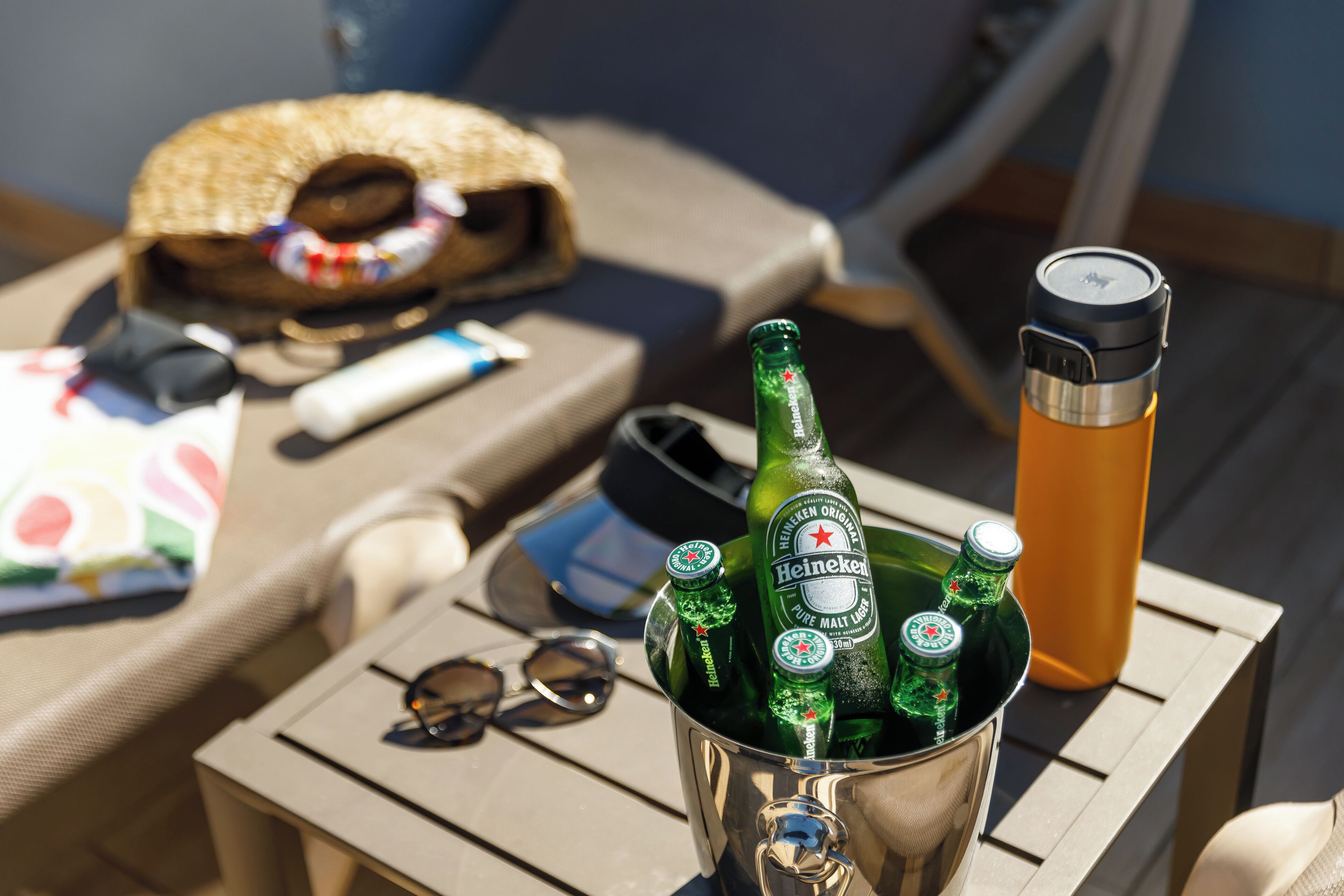 Un balde metálico sobre una mesa con botellas de cerveza Heineken, una botella de agua y gafas de sol.