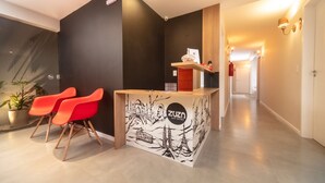 A reception area with a counter, two red chairs, a potted plant, and a wall with a cityscape illustration. - Zuza Slim Suítes (Maceió)