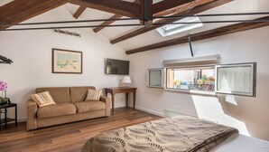 Une pièce avec un plafond en bois, un lit recouvert d’une couvre-lit à motifs, un canapé avec des coussins, un téléviseur fixé au mur et une fenêtre avec des stores.