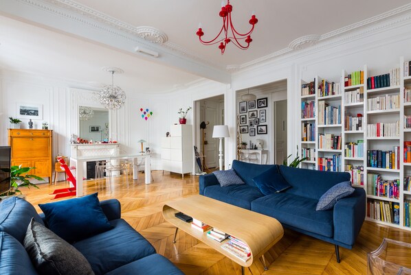 Een ruime woonkamer met een blauwe bank, een houten salontafel en een boekenplank vol boeken.