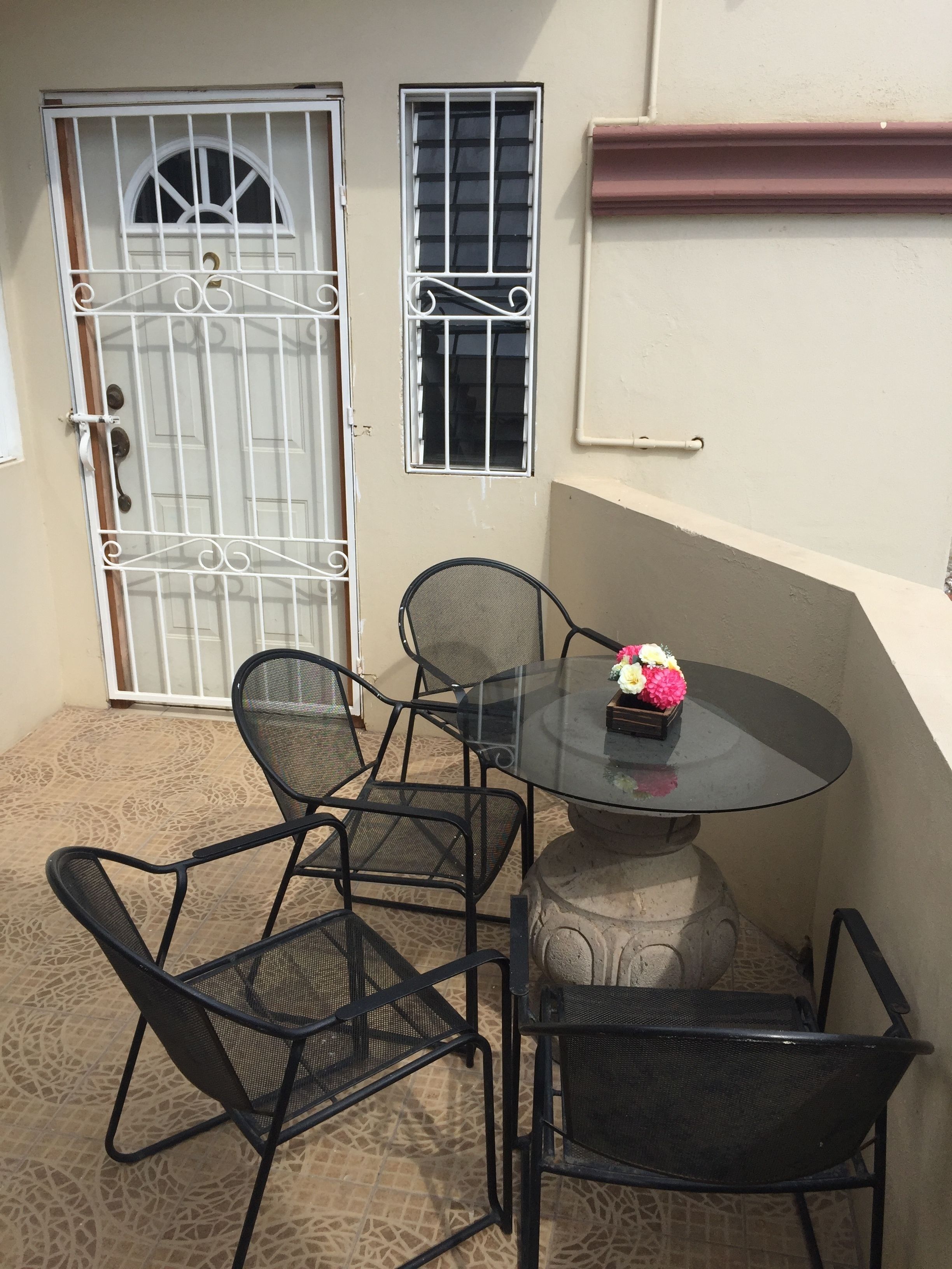 A small outdoor seating area with a round table and four chairs, a decorative vase with flowers, and a barred gate.