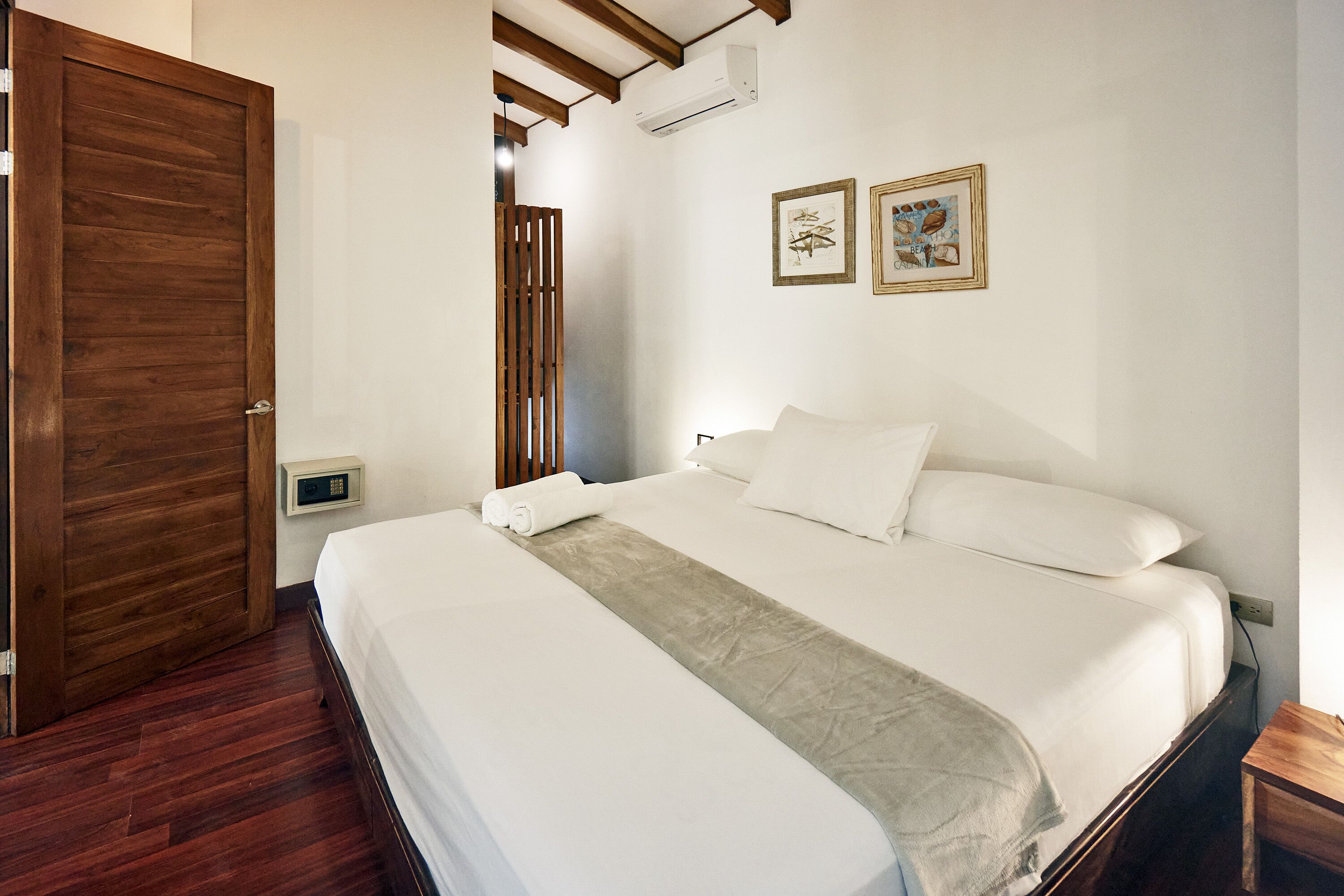A hotel room with a wooden door, a bed with white linens, a beige throw blanket, and two framed pictures on the wall.