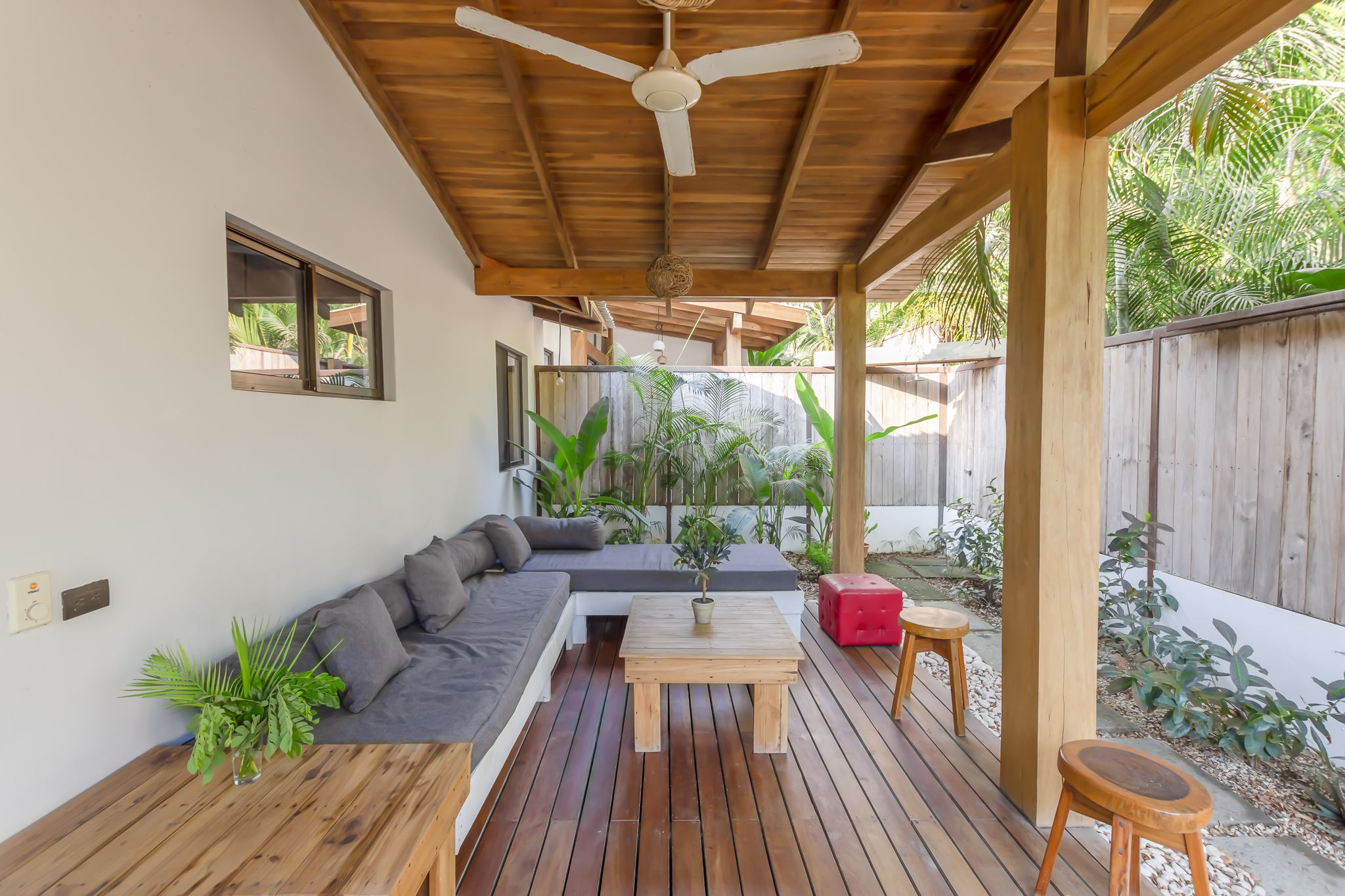 A covered outdoor seating area with a wooden ceiling fan, a sofa, a small table, and a potted plant.