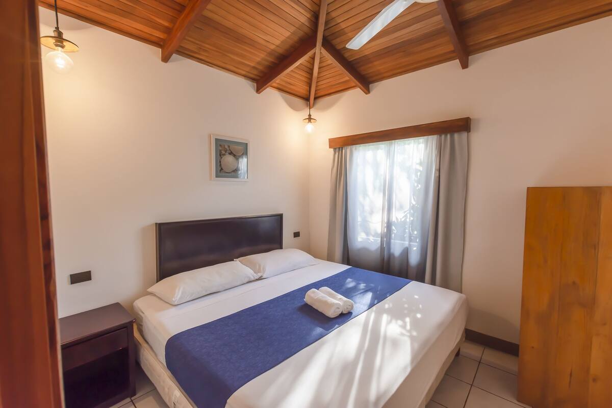 A neatly made bed with a blue bedspread and white pillows, a wooden headboard, a nightstand, a ceiling fan, and a window with curtains.