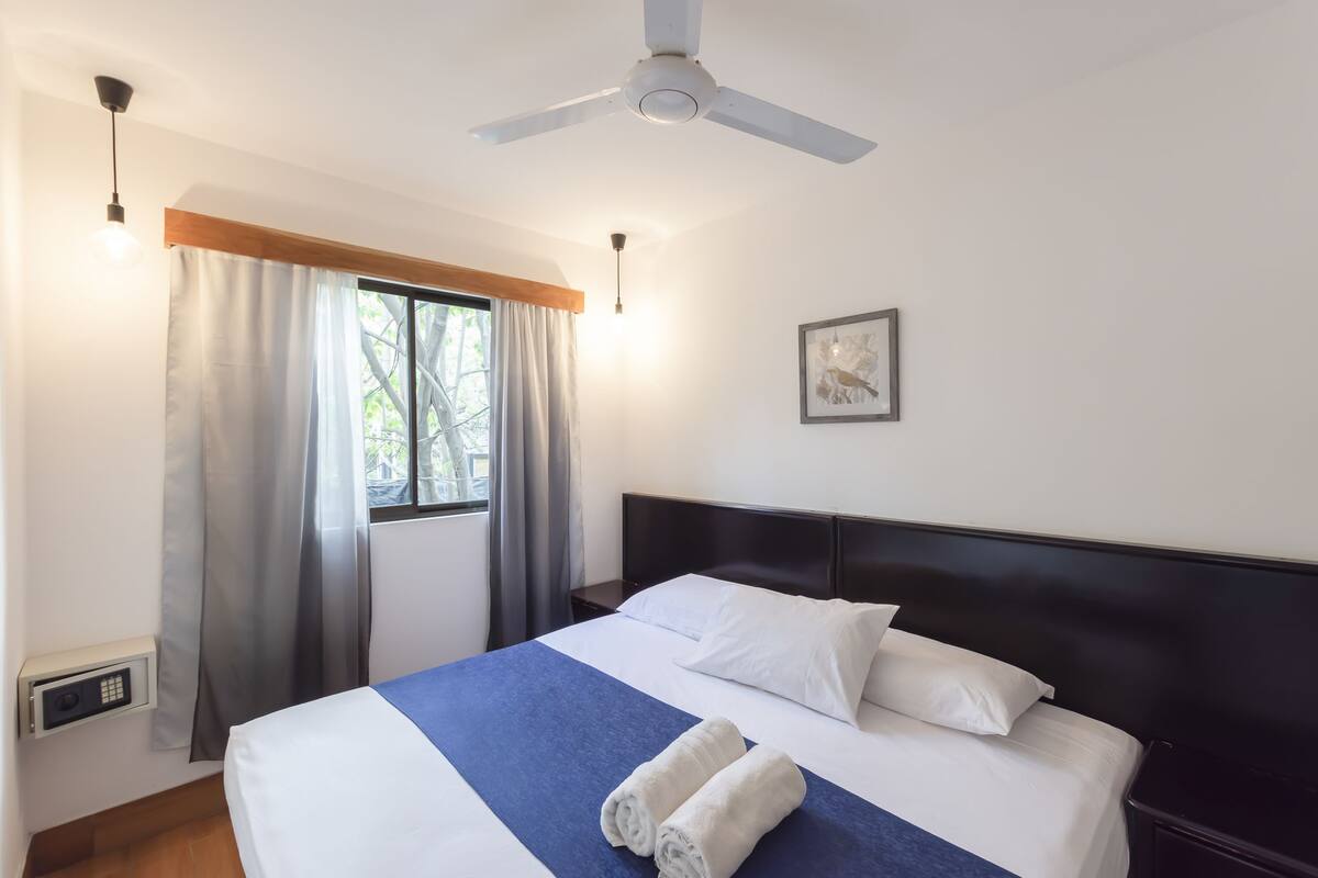 A neatly made bed with white and blue bedding, a ceiling fan, a window with curtains, and a framed picture on the wall.