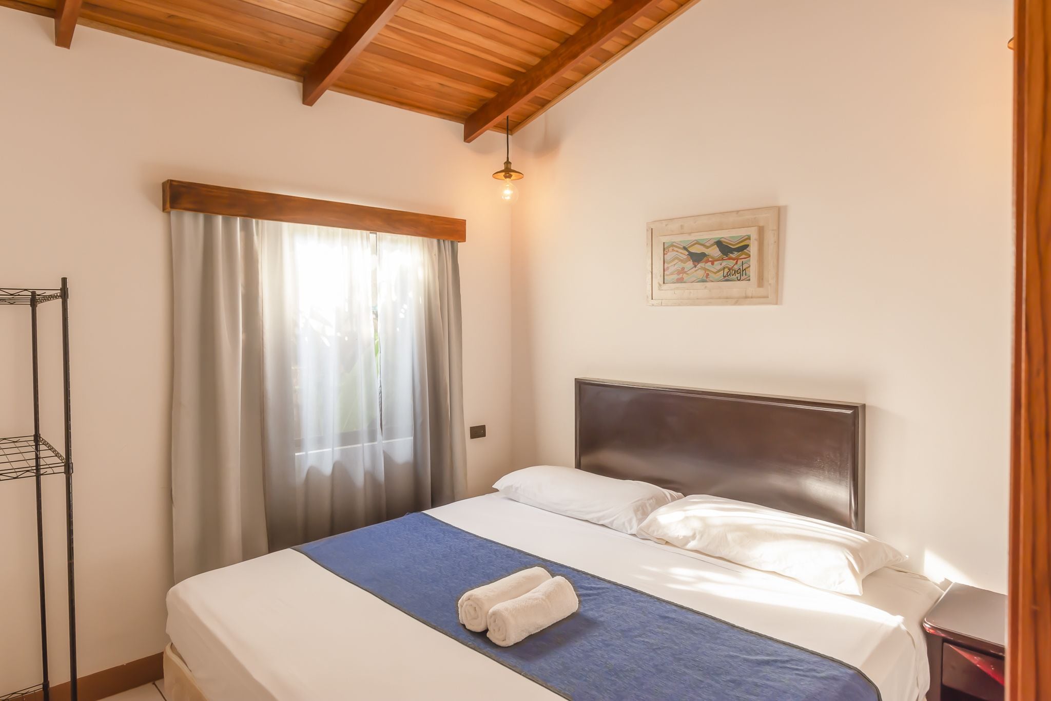 A neatly made bed with a blue bedspread and white pillows, a wooden headboard, a hanging light fixture, and a painting on the wall.