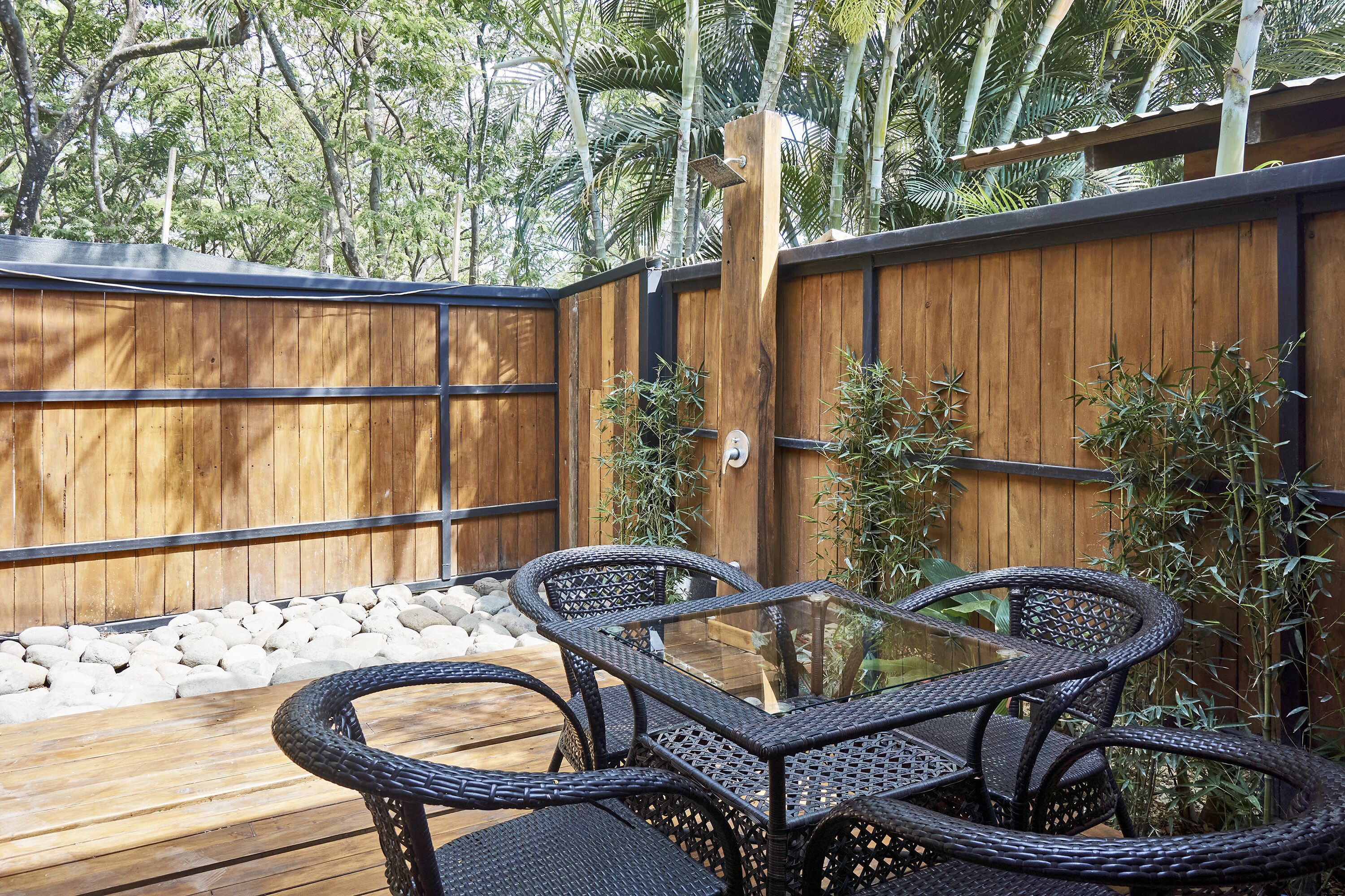 Een houten terras met rieten stoelen en een glazen tafel, omringd door een houten hek en weelderige begroeiing.