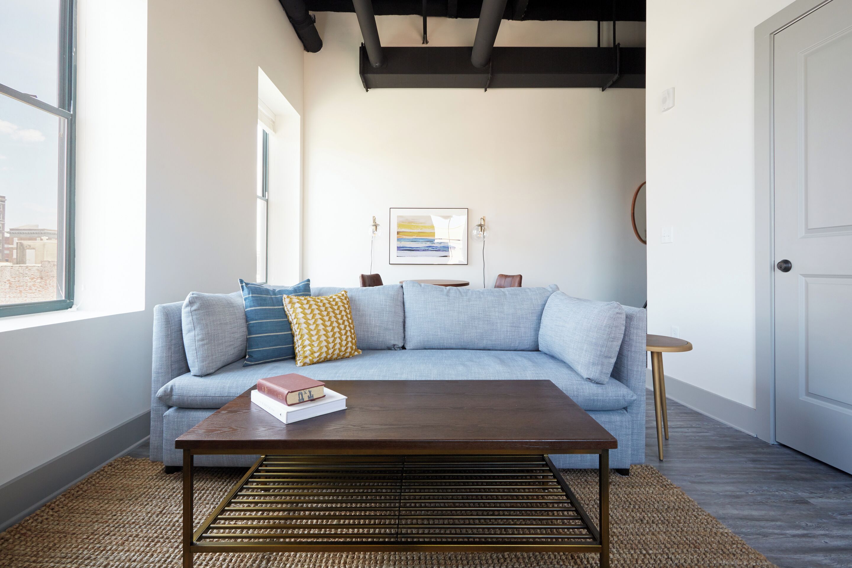 A modern living room with a light blue sofa, a wooden coffee table, and a large window providing a view of the city.