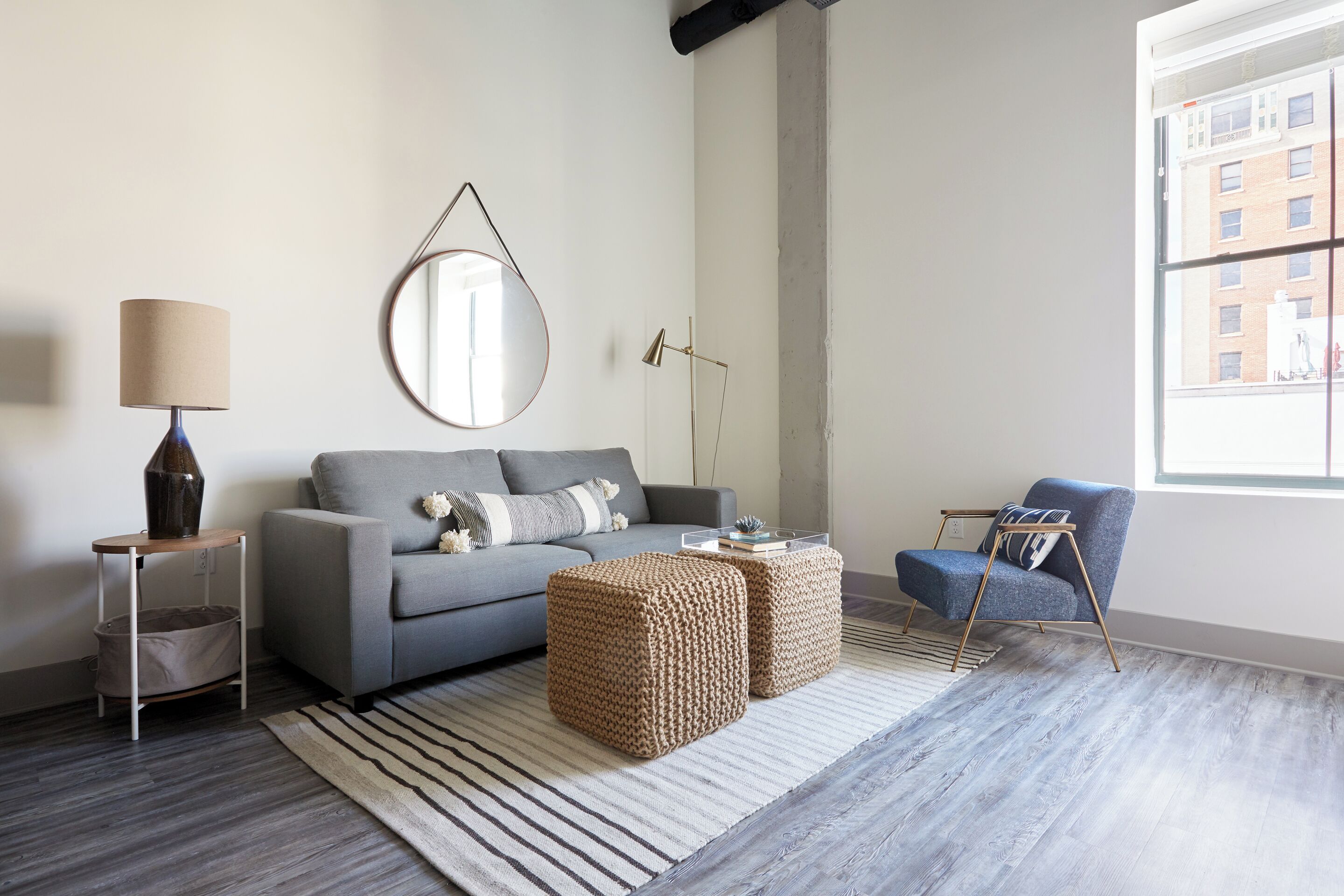 A modern living room with a grey sofa, a round mirror, a blue armchair, and two woven ottomans.