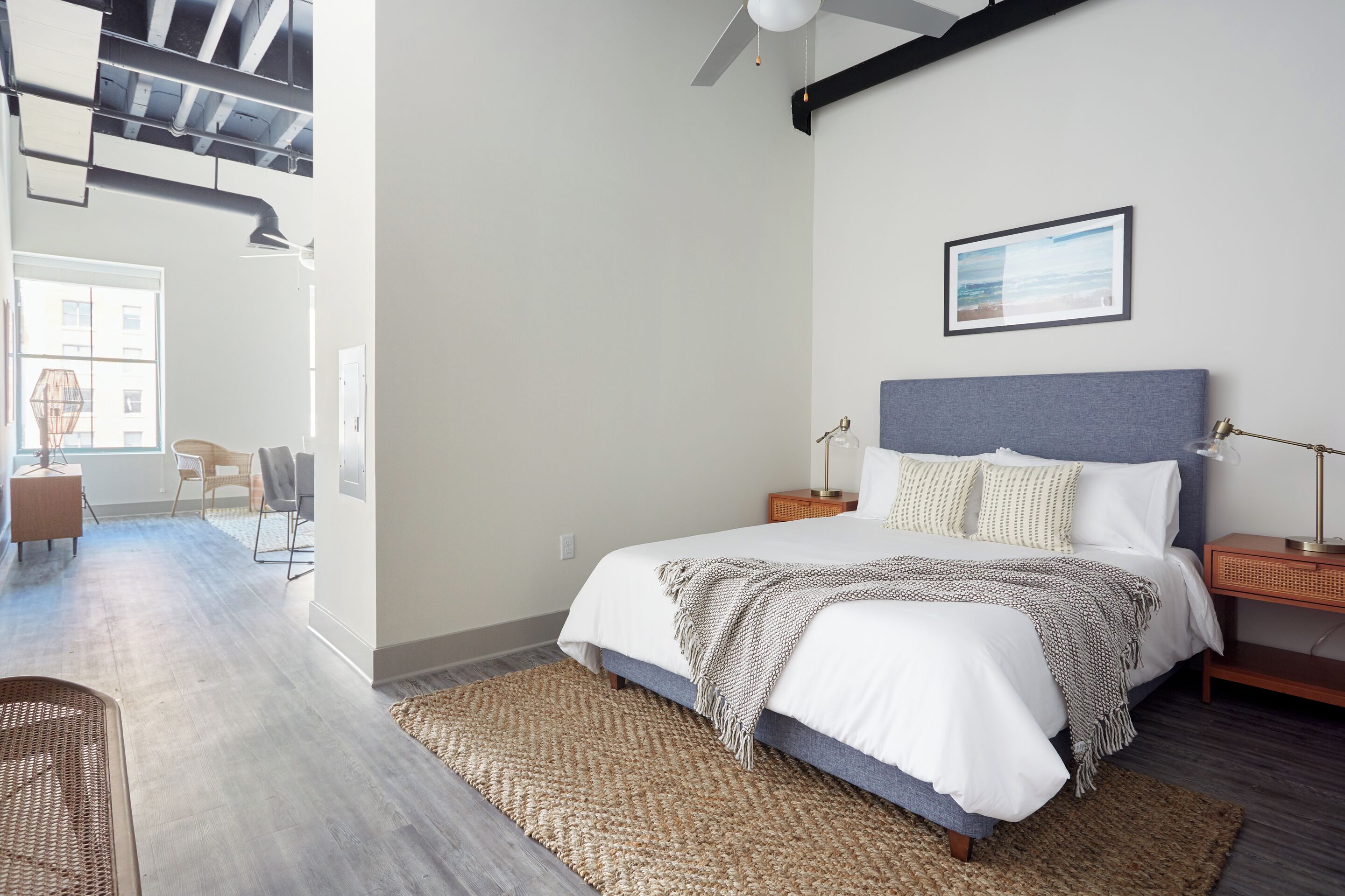 A modern bedroom with a large bed, a ceiling fan, a framed picture on the wall, and a view of the outdoors through a window.