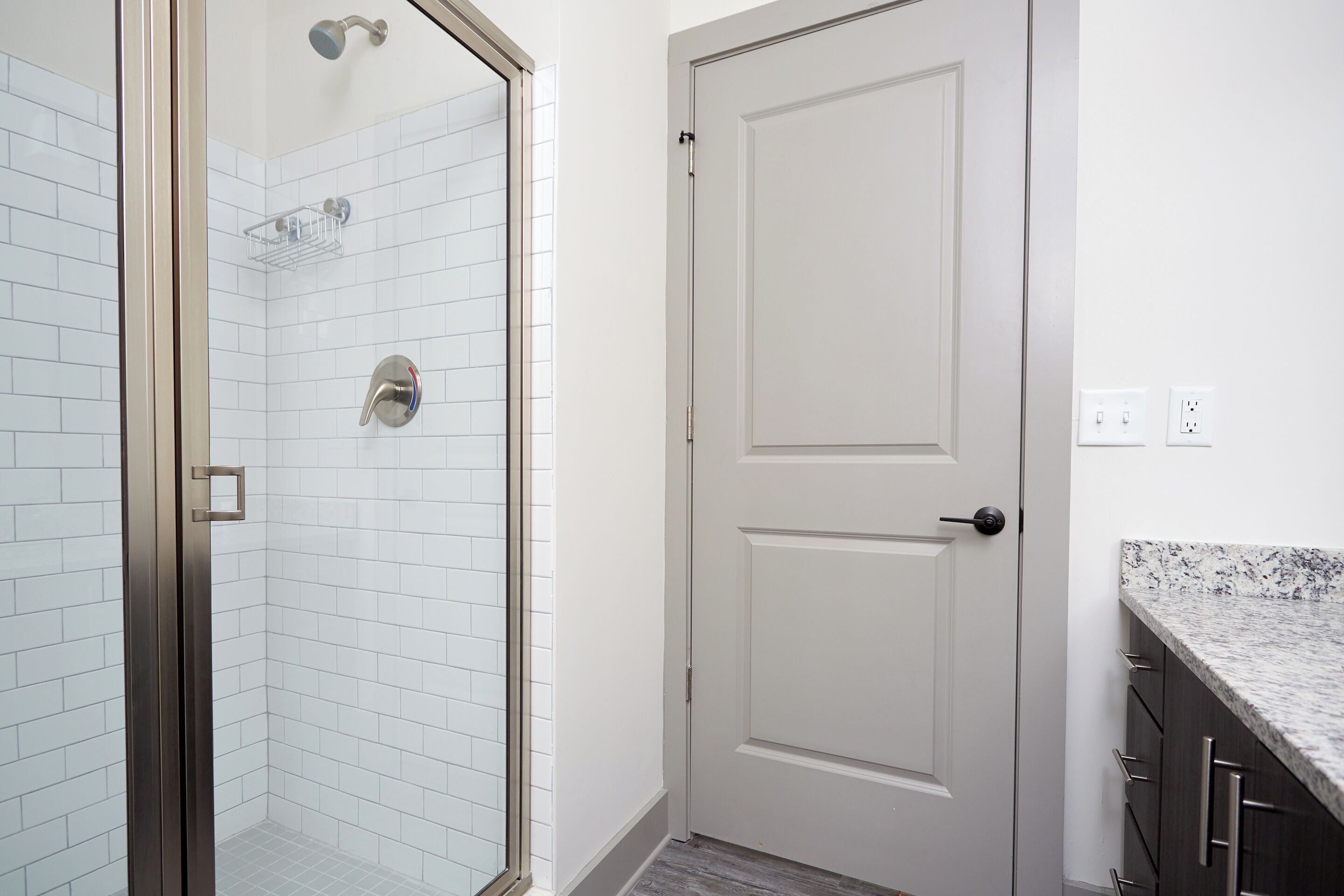 Un bagno con piastrelle bianche tipo metro, box doccia in vetro, porta bianca e piano di lavoro in granito.