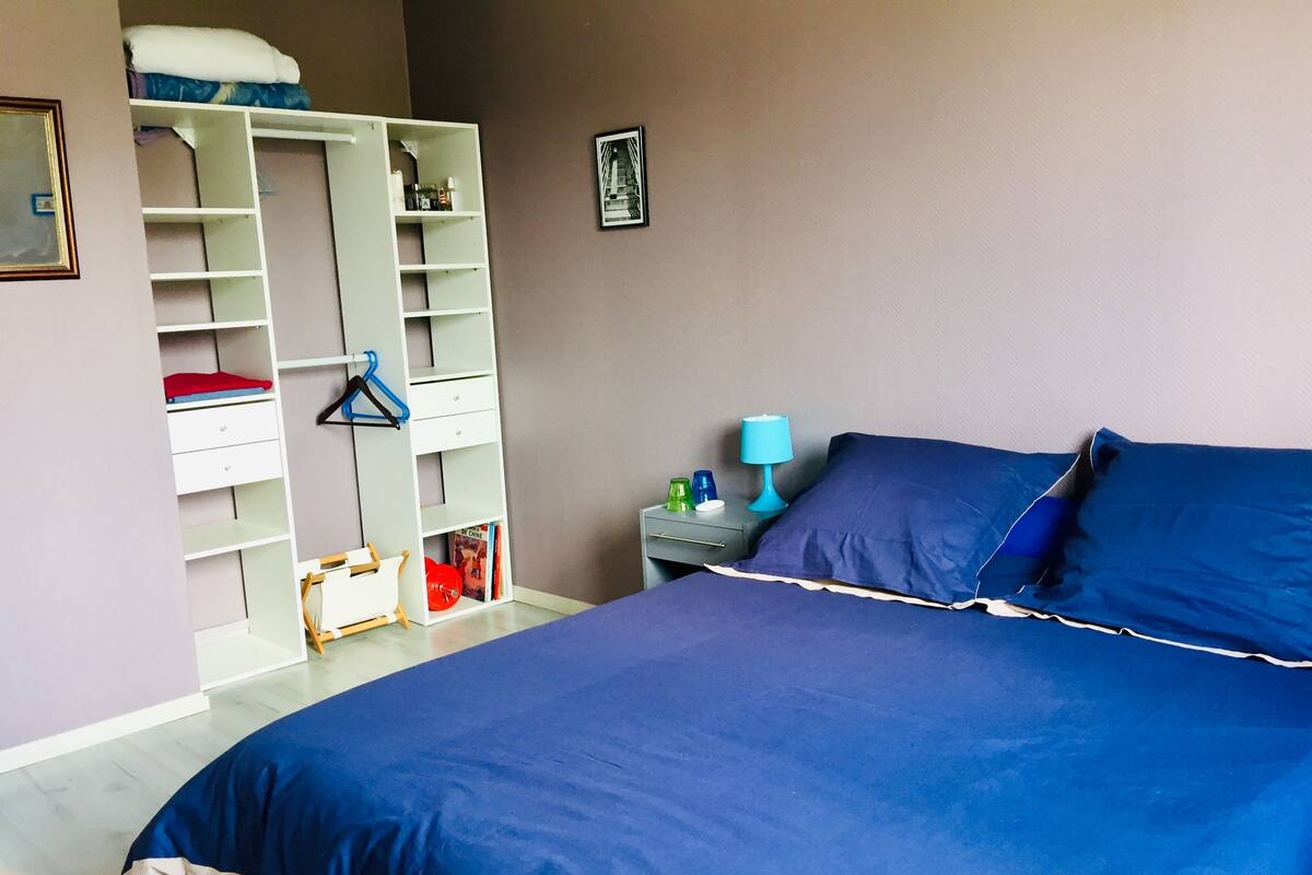 A bedroom with a large bed, a wardrobe, a nightstand, and a wall-mounted shelf.
