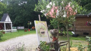 A rustic outdoor setting with a wooden fence, a tree, and a sign with a cartoon character. - Jolie halte normande (Balines)