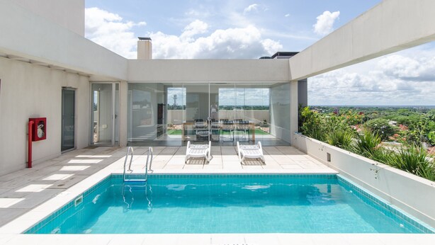A rooftop swimming pool with clear blue water, surrounded by white walls and a glass door leading to a balcony with a view of the city. - OXO Bertoni Luxury in Villamorra (Asunción)