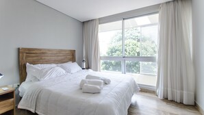 A modern bedroom with a large bed, a wooden headboard, and a view of greenery through the window. - OXO Bertoni Luxury in Villamorra (Asunción)