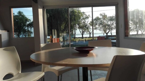 A modern dining area with a round table and chairs, large windows with a view of trees and a body of water. - Alojamiento La Roja (Chascomus)