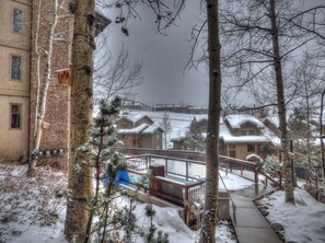 A snowy outdoor area with a wooden deck, a metal railing, and a building with snow-covered roofs in the background. - Lift Tickets Ski To Your Door, Wifi, Deck, Hot Tub (Steamboat Springs)