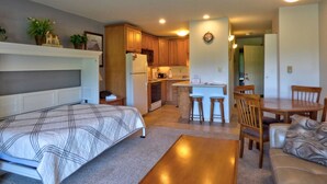 A compact living space with a bed, a dining table with chairs, a kitchen area with wooden cabinets, and a small island with stools. - Lift Tickets Heart Of The Base with Private Deck & Fireplace (Steamboat Springs)