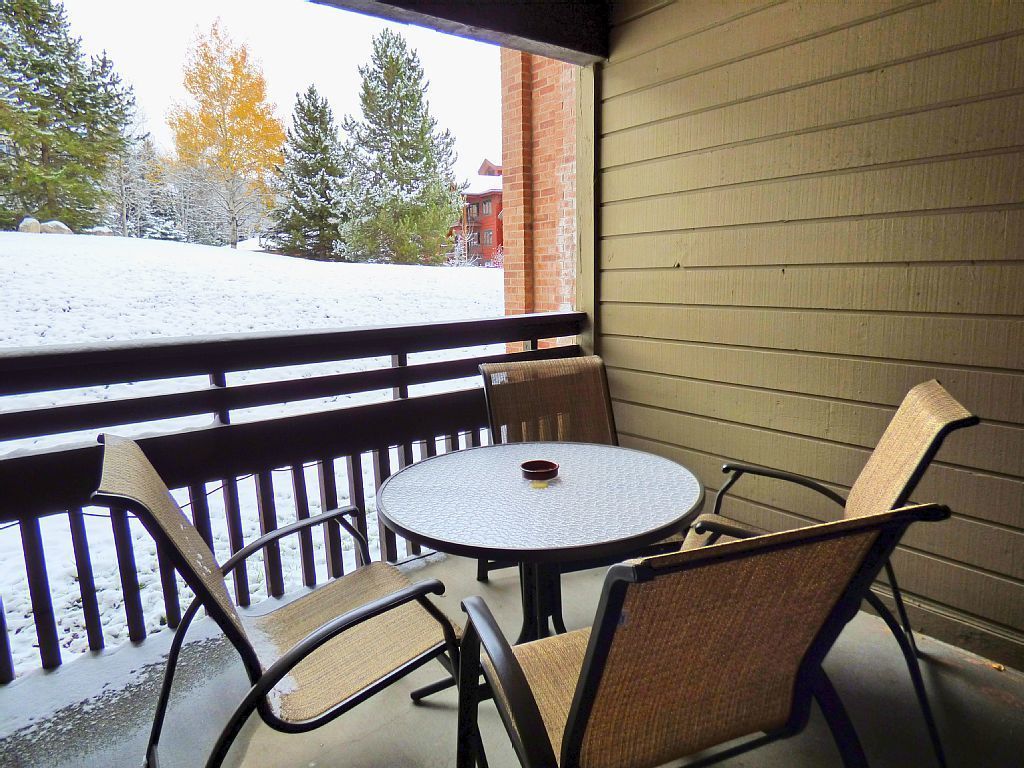 Un espace extérieur aménagé avec une zone de repos couverte, comprenant une table ronde et quatre chaises, offrant une vue sur un paysage enneigé avec des arbres et un bâtiment en arrière-plan.