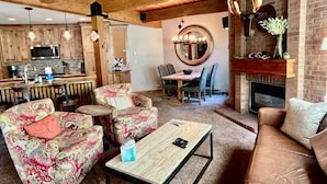 Un salon chaleureux avec une cheminée, une table à manger et une cuisine en arrière-plan.