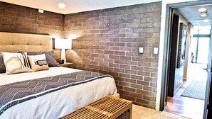 A modern hotel room with a brick accent wall, a large bed with patterned bedding, a wooden bench, and a visible door leading to a hallway. - Lift Tickets Perfect Penthouse Rustic Finishes Fireplace (Steamboat Springs)