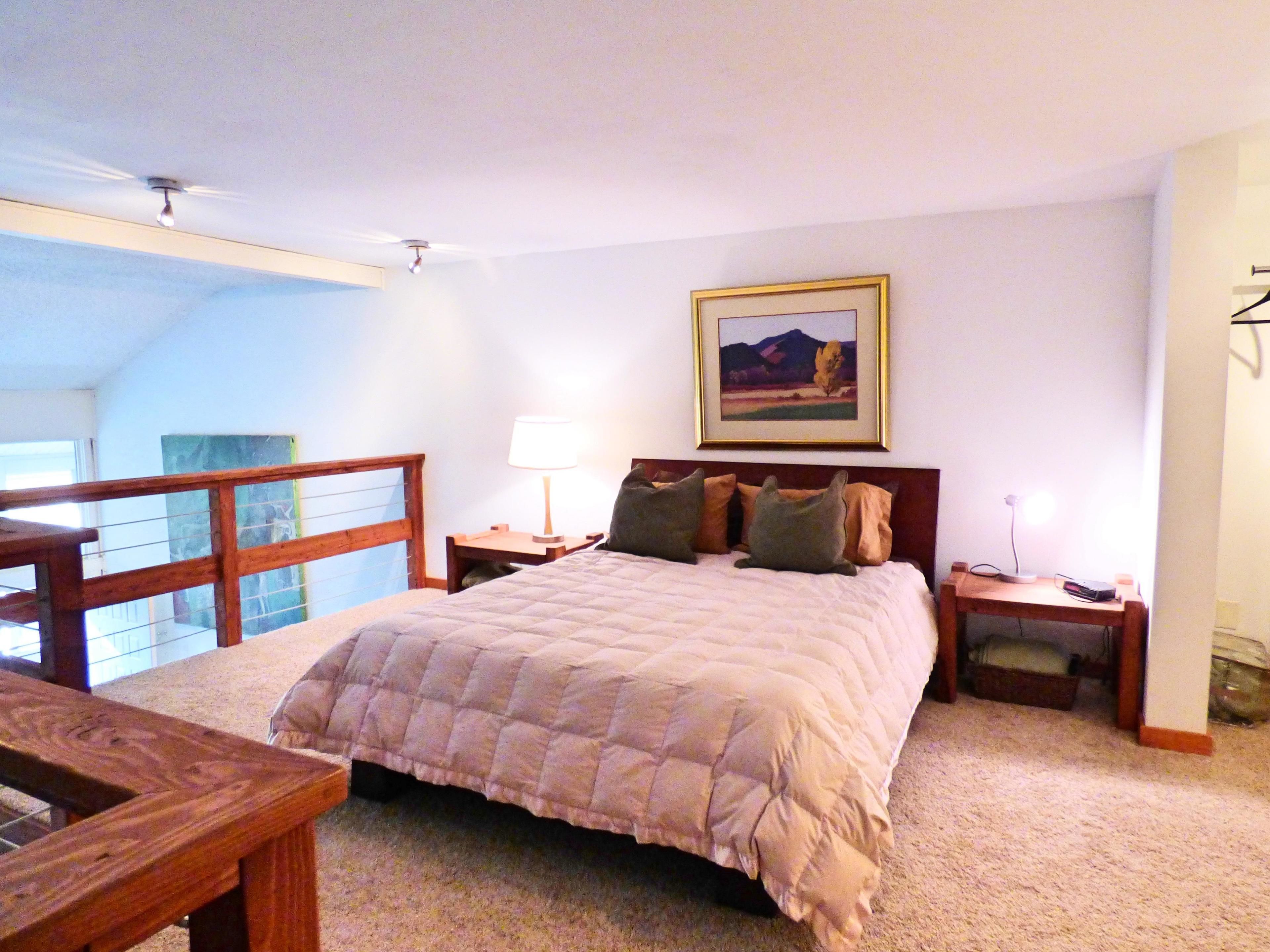 A bedroom with a large bed, a desk, a chair, a nightstand, a lamp, and a framed picture on the wall.