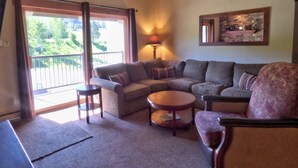 A living room with a sectional sofa, a round coffee table, and a patterned armchair. There is a balcony with a railing and a lamp on a side table. - Lift Tickets Newly Remodeled Sleeps 8 Pool, Hot Tubs,fireplace, Private Deck (Steamboat Springs)