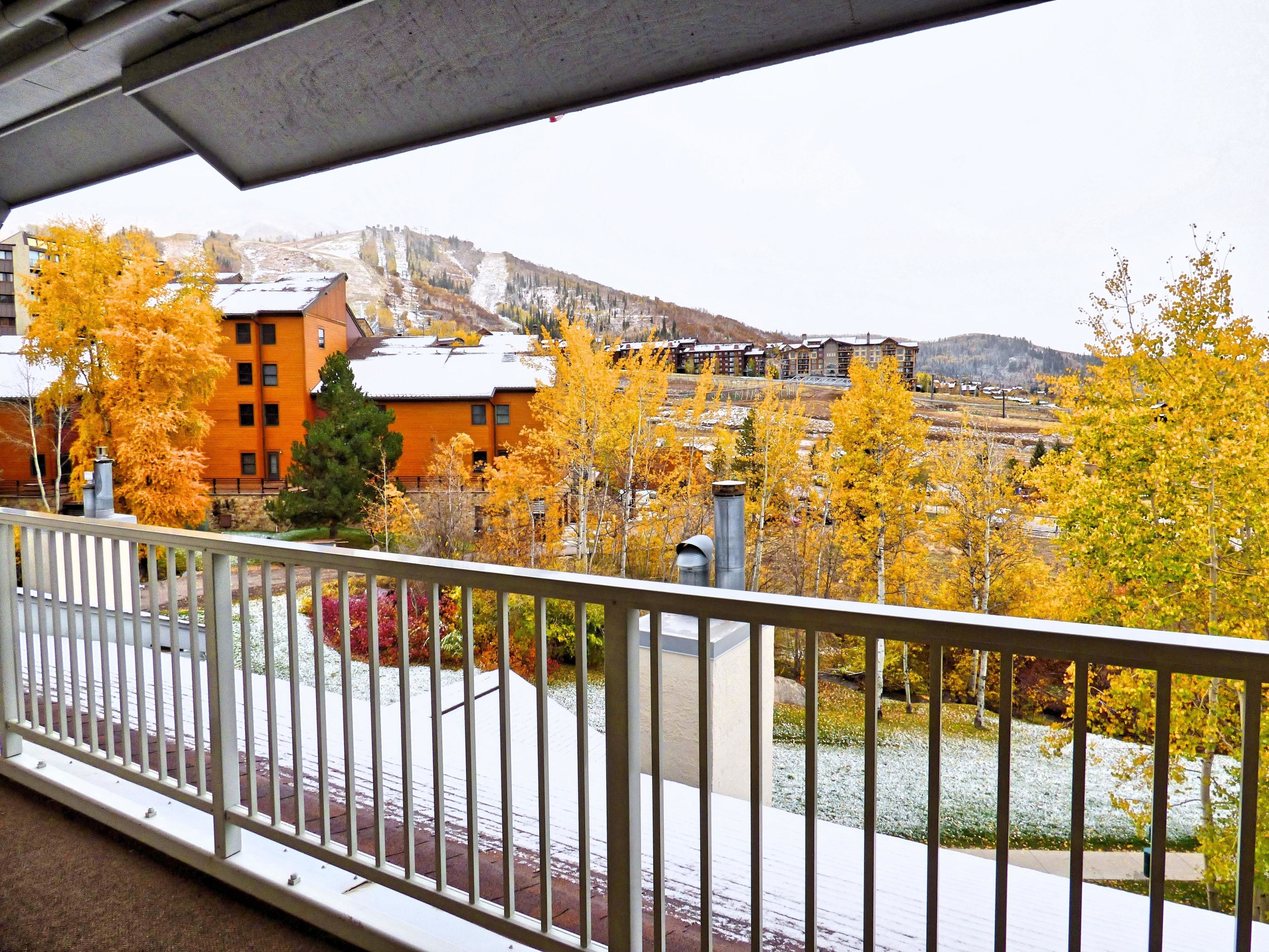 A balcony with a view of a building with orange siding, a snow-covered landscape, and autumn-colored trees.