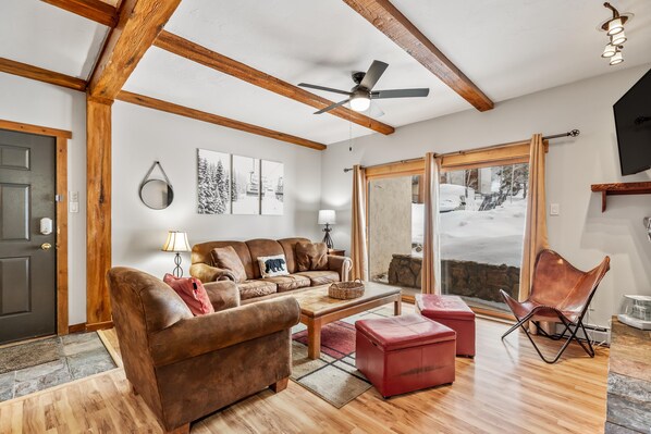 A cozy living room with a brown leather sofa, a red ottoman, and a wooden coffee table. There is a ceiling fan, a large window with a view of a snowy landscape, and a mounted TV.