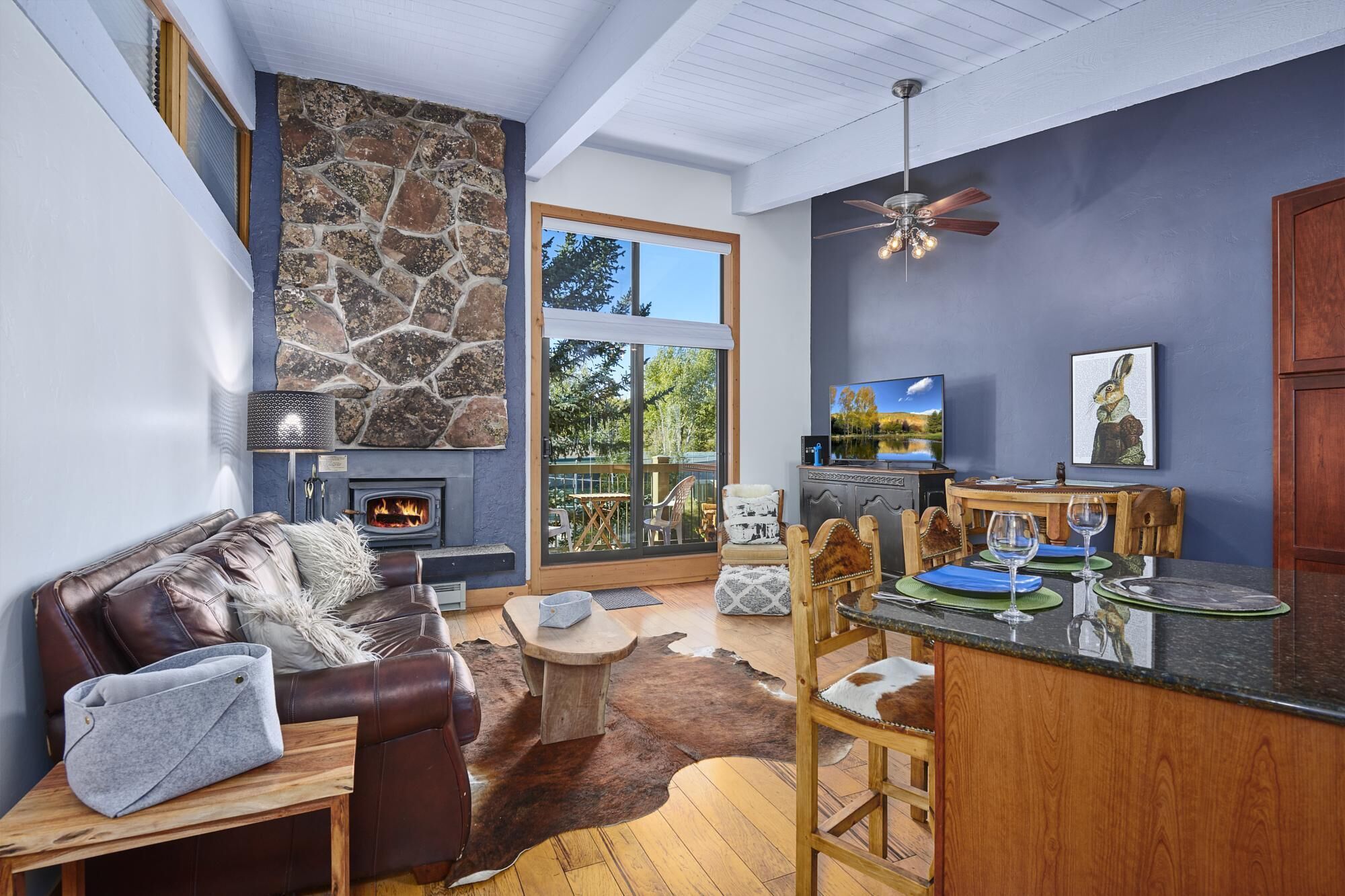 A cozy living room with a stone fireplace, leather sofa, and a dining area with wooden chairs and a glass-top table.