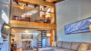 A spacious living room with a high ceiling, a fireplace, a staircase, and a large window with a mountain view. - Lift Tickets Pool, Hot Tubs, Shuttle & Private Deck Mountain Village (Steamboat Springs)