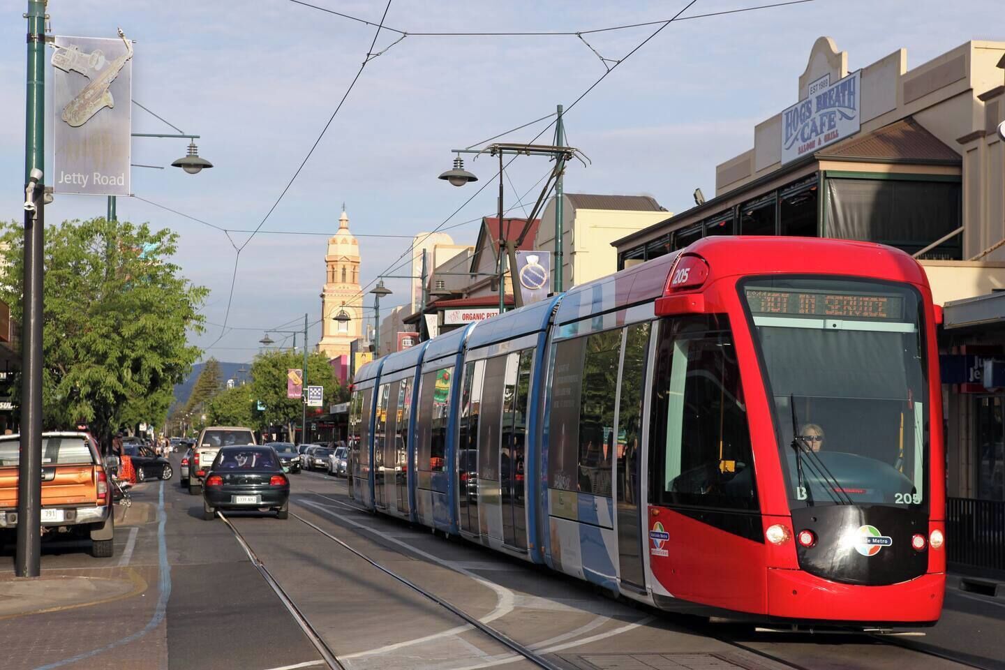 Eine Straßenbahn fährt auf einer Stadtstraße, im Hintergrund sind Gebäude und ein Turm zu sehen.