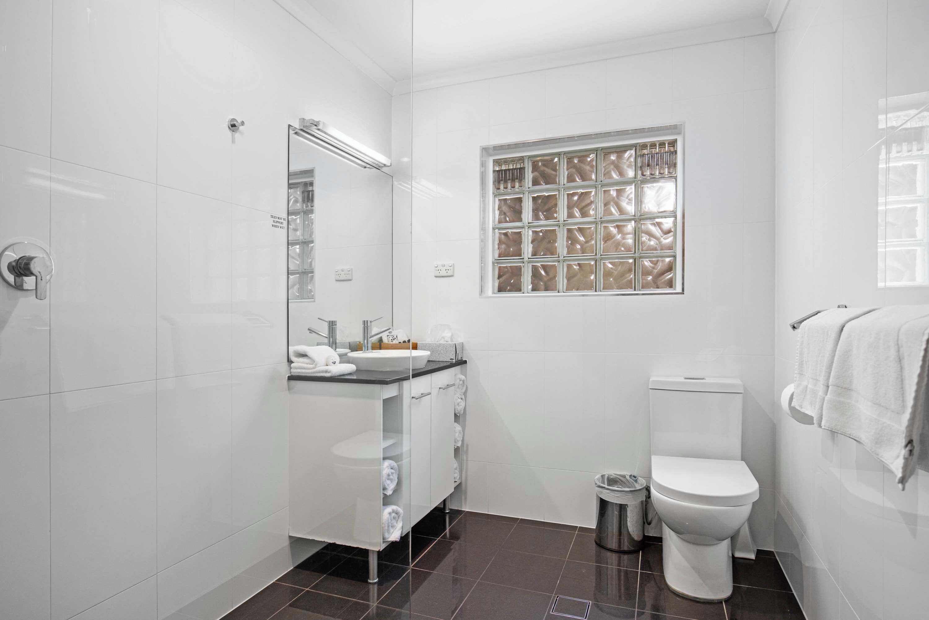 Une salle de bain moderne avec une douche à parois de verre, un lavabo blanc avec un plan de travail, des toilettes et une fenêtre ornée d’un motif décoratif.