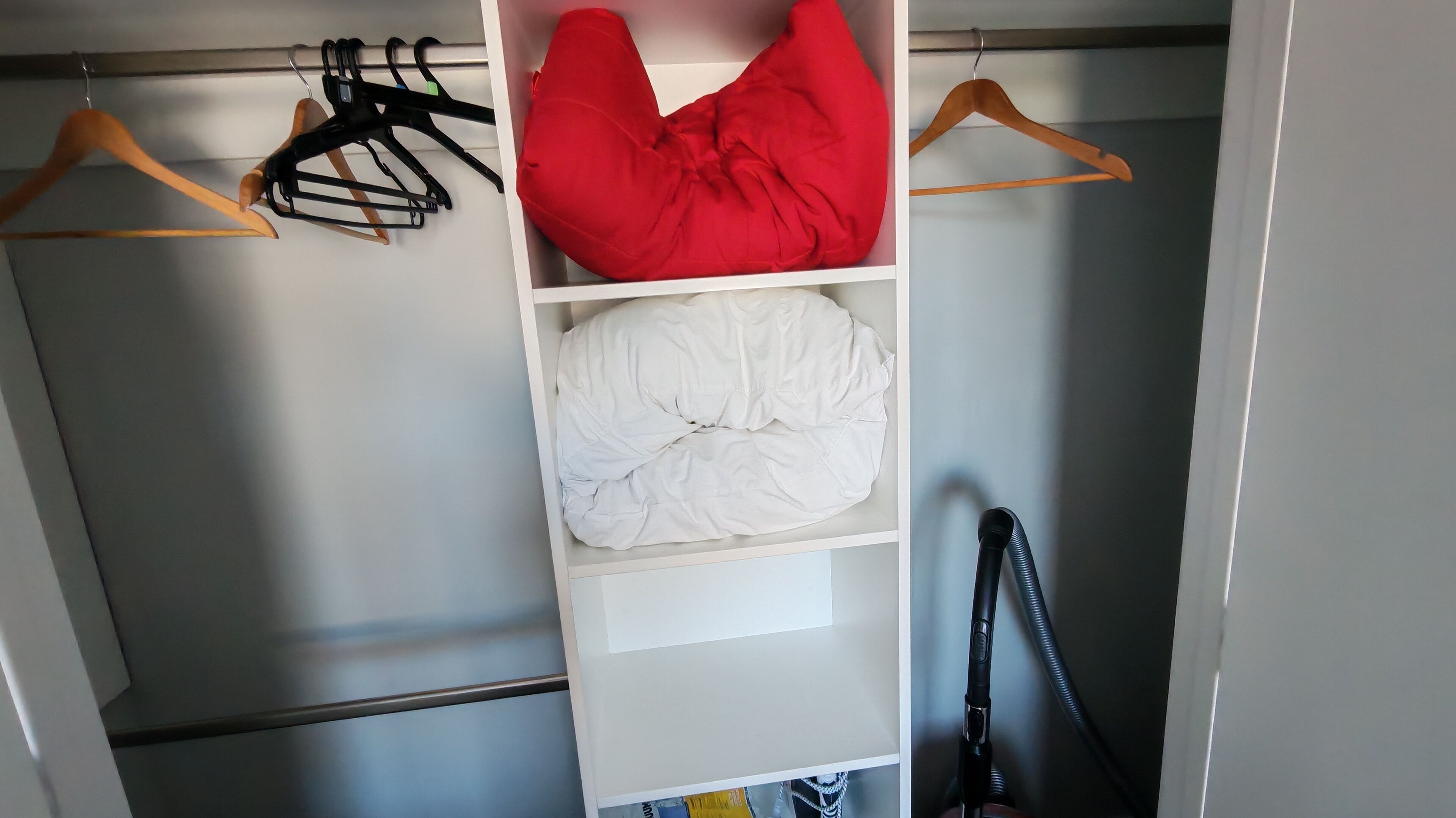 A closet with hanging clothes, a red cushion, and a vacuum cleaner.