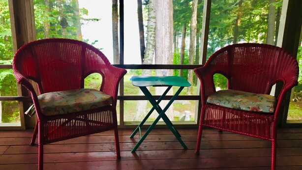 Deux fauteuils en osier ornés de coussins à motifs floraux, placés sur une terrasse en bois, une petite table et une vue sur une forêt, visible à travers de grandes fenêtres.
