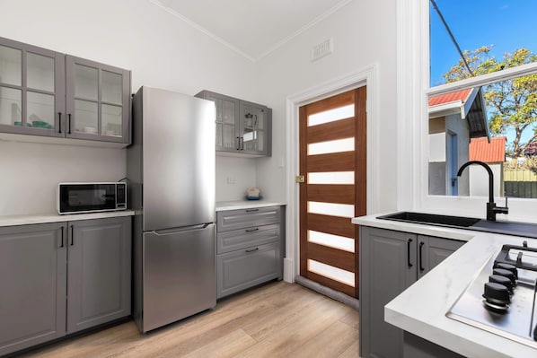 A modern kitchen with grey cabinets, a stainless steel refrigerator, a microwave, a sink, and a window with a view of a house.