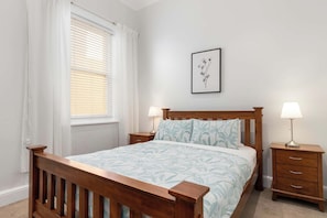 A bedroom with a wooden bed, two bedside tables with lamps, a window with blinds, and a framed picture on the wall. - Cosy Glenelg South Family Cottage| Close To Beach | Pets Welcome (Glenelg South)