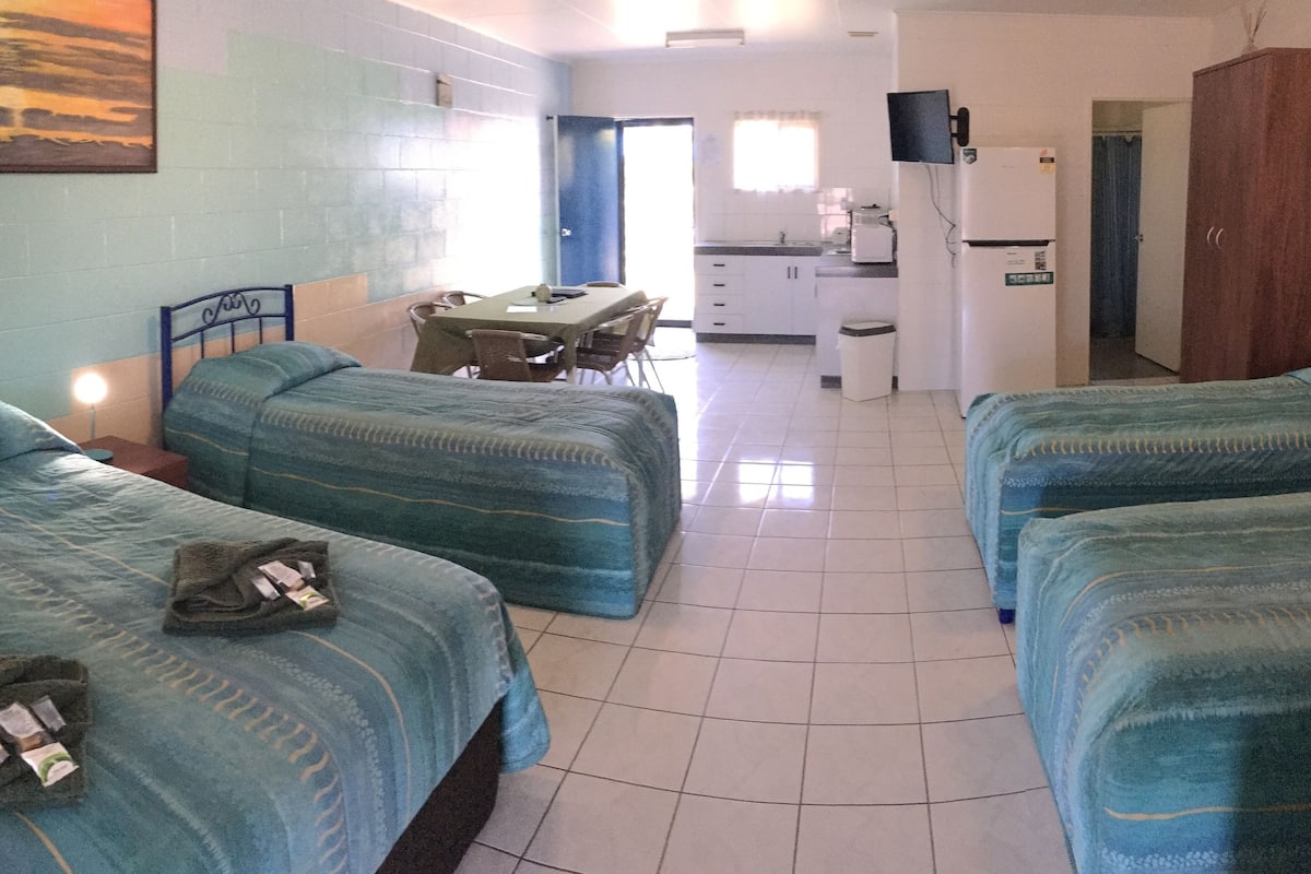 Una habitación de hotel con tres camas, un escritorio, una silla, un televisor y una kitchenette.