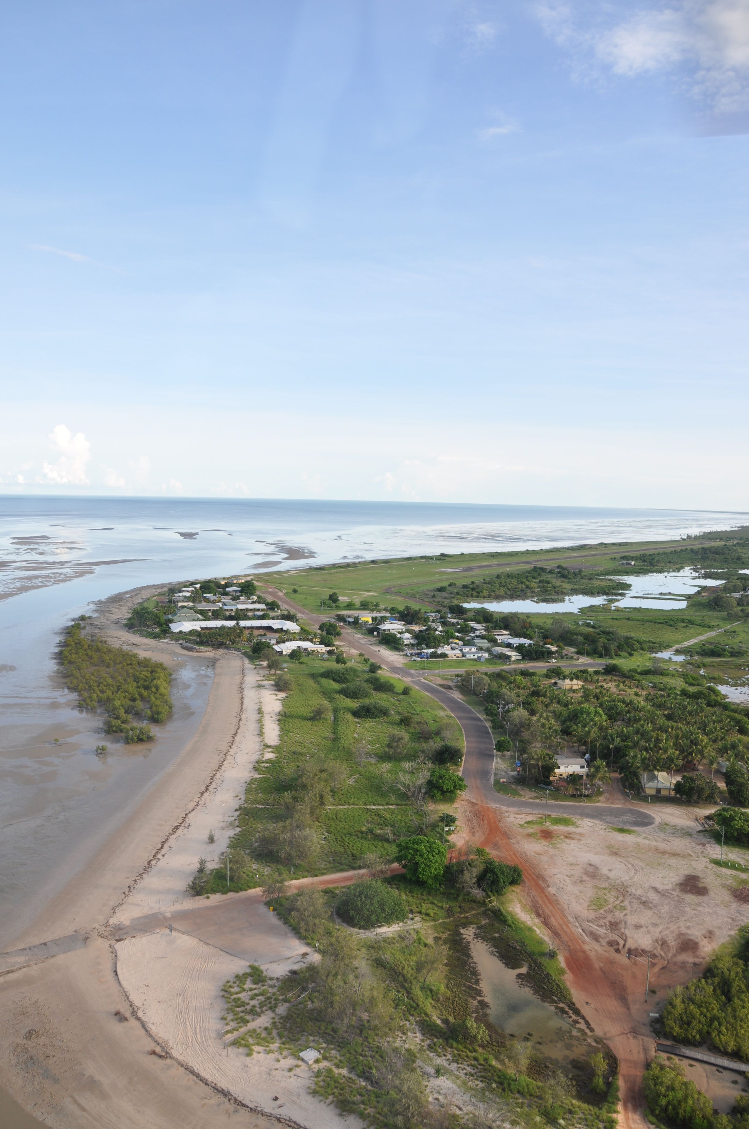 Luftaufnahme einer Küstenlandschaft mit Strand, einer Straße und vereinzelten Gebäuden.