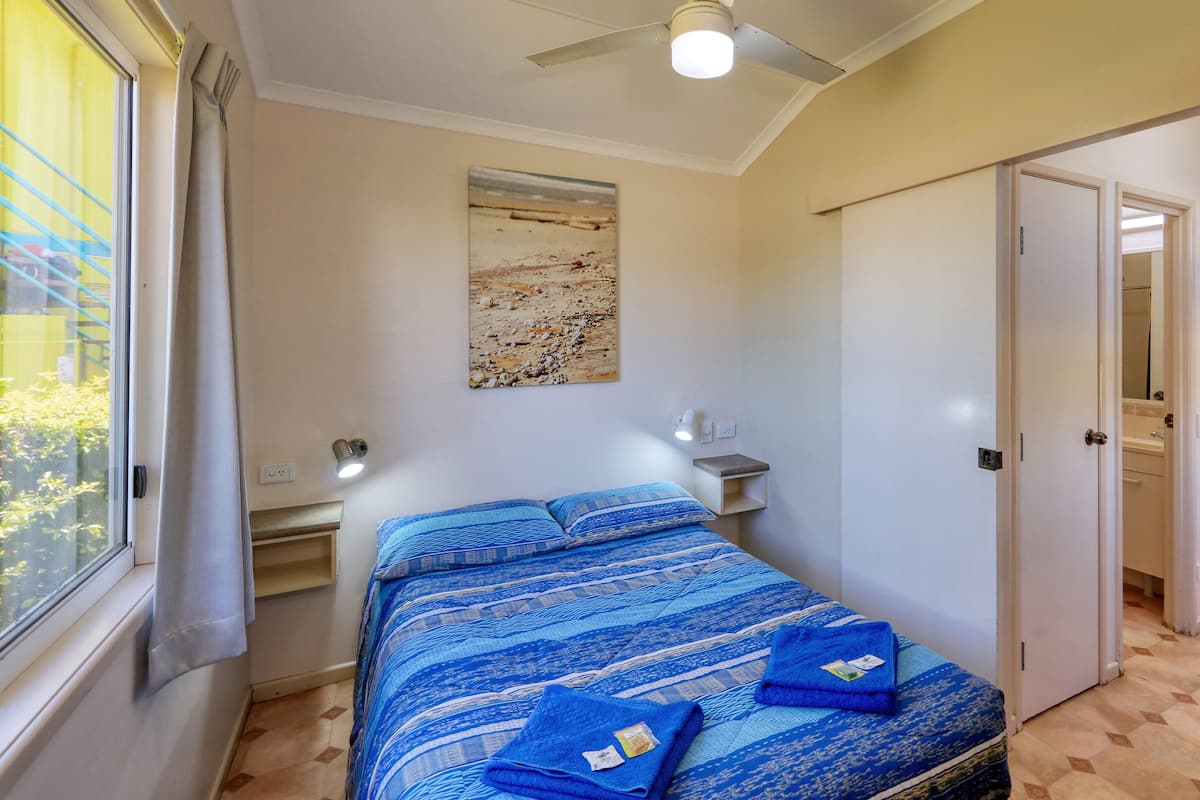 Un dormitorio con cama, ventilador de techo, mesita de noche, un cuadro y puerta al baño.