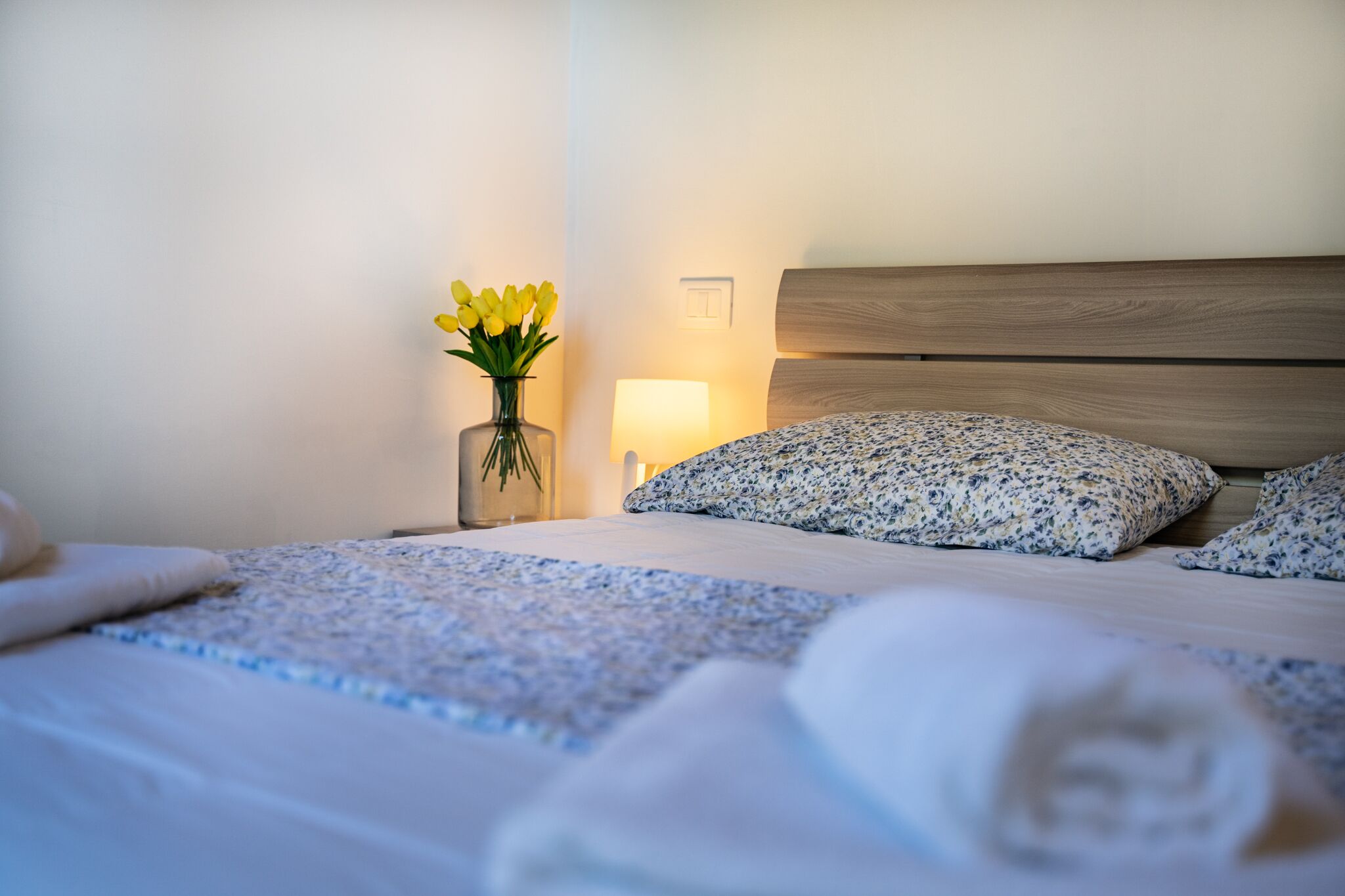 Una cama bien hecha con ropa de cama floral, una lámpara de noche y un jarrón con flores amarillas.