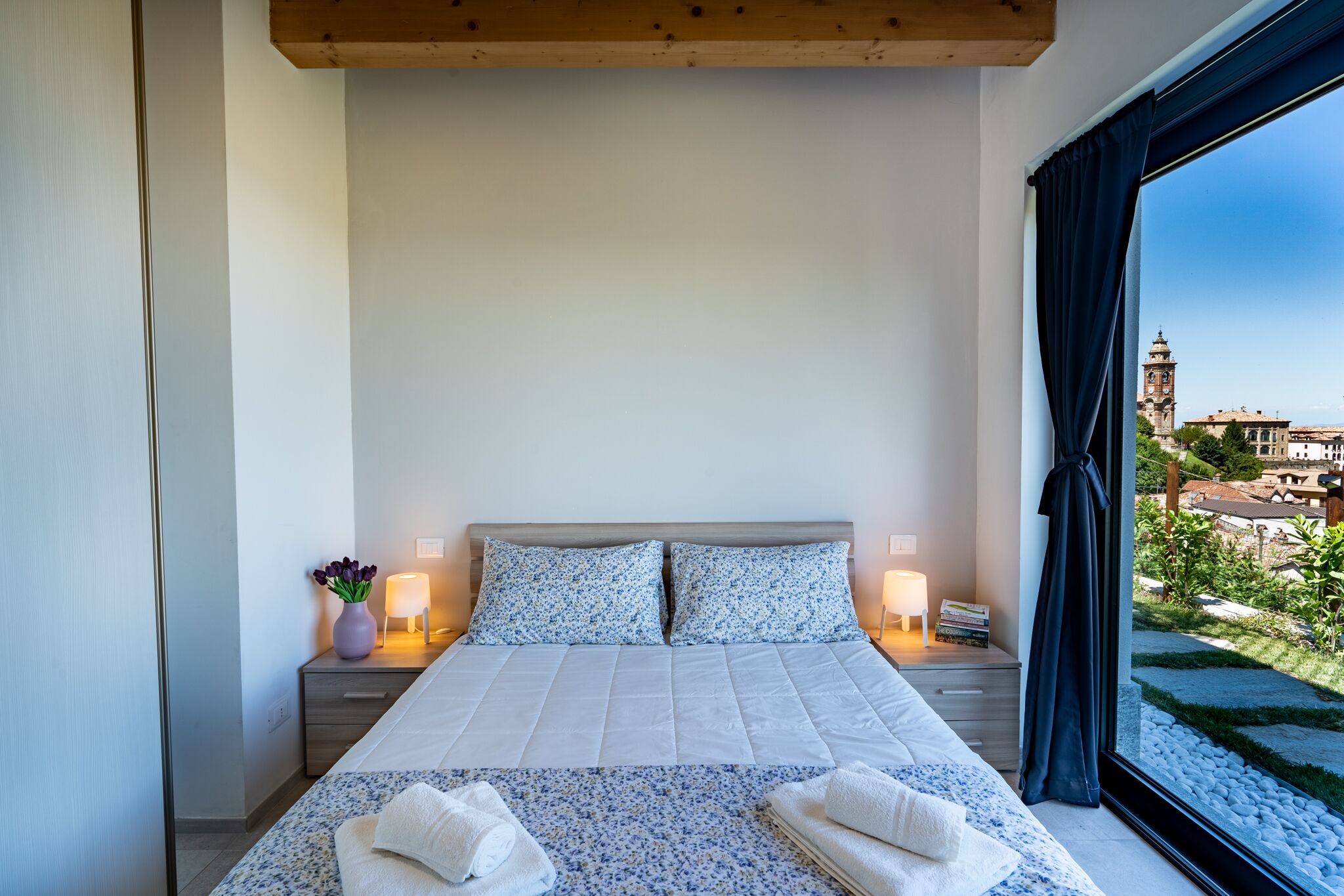A modern hotel room with a large bed, two bedside tables, a nightstand, a window with curtains, and a view of a cityscape.