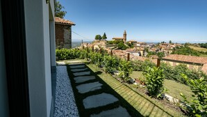 A balcony with a stone pathway leading to a lush garden, overlooking a historic village with traditional buildings and a prominent tower. - ONE BEDROOM HOLIDAY FLAT WITH VIEW (Diano d'Alba)