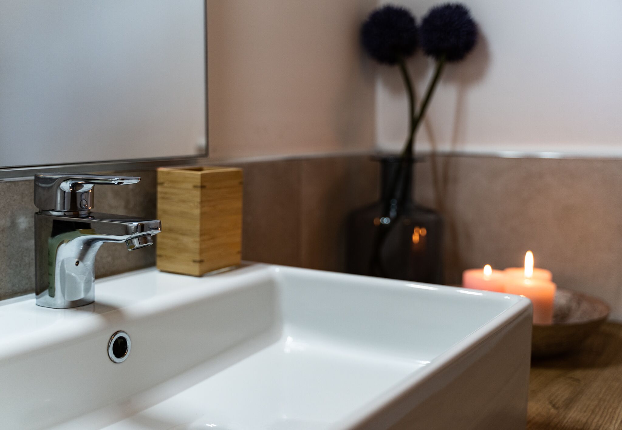 A white bathroom sink with a chrome faucet, a wooden soap dish, and a lit candle in the background.