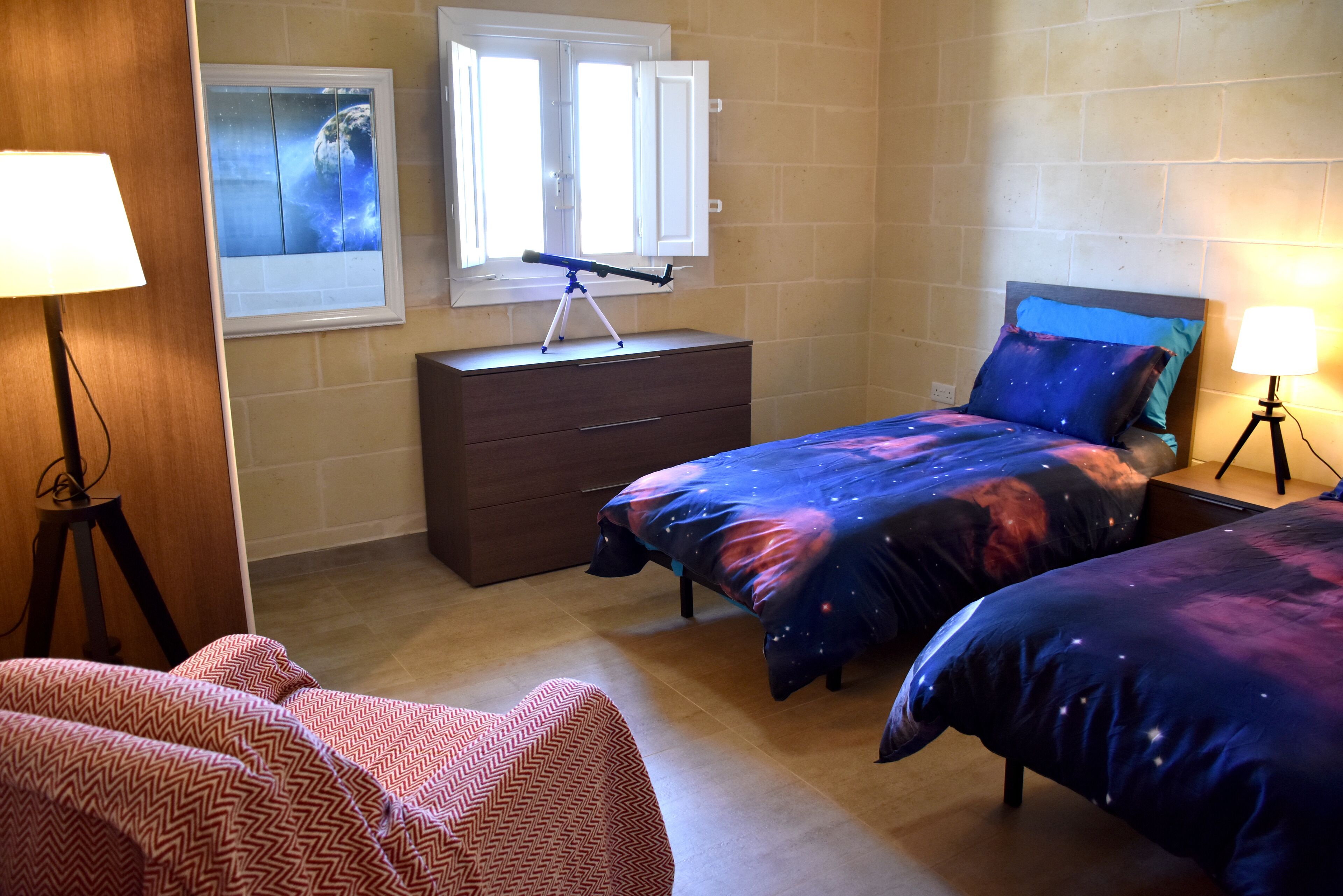 A bedroom with two beds, a dresser, a window with curtains, and a lamp.