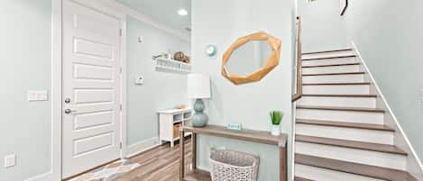 A hallway with a wooden floor, a white door, a console table with a lamp, a wicker basket, and a staircase with wooden steps.