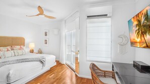 A modern bedroom with a bed, a ceiling fan, a wall-mounted air conditioner, a TV displaying a sunset scene, and a wicker chair.