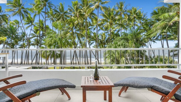 A balcony with two wooden chairs and a small table, overlooking a beach with palm trees and the ocean.