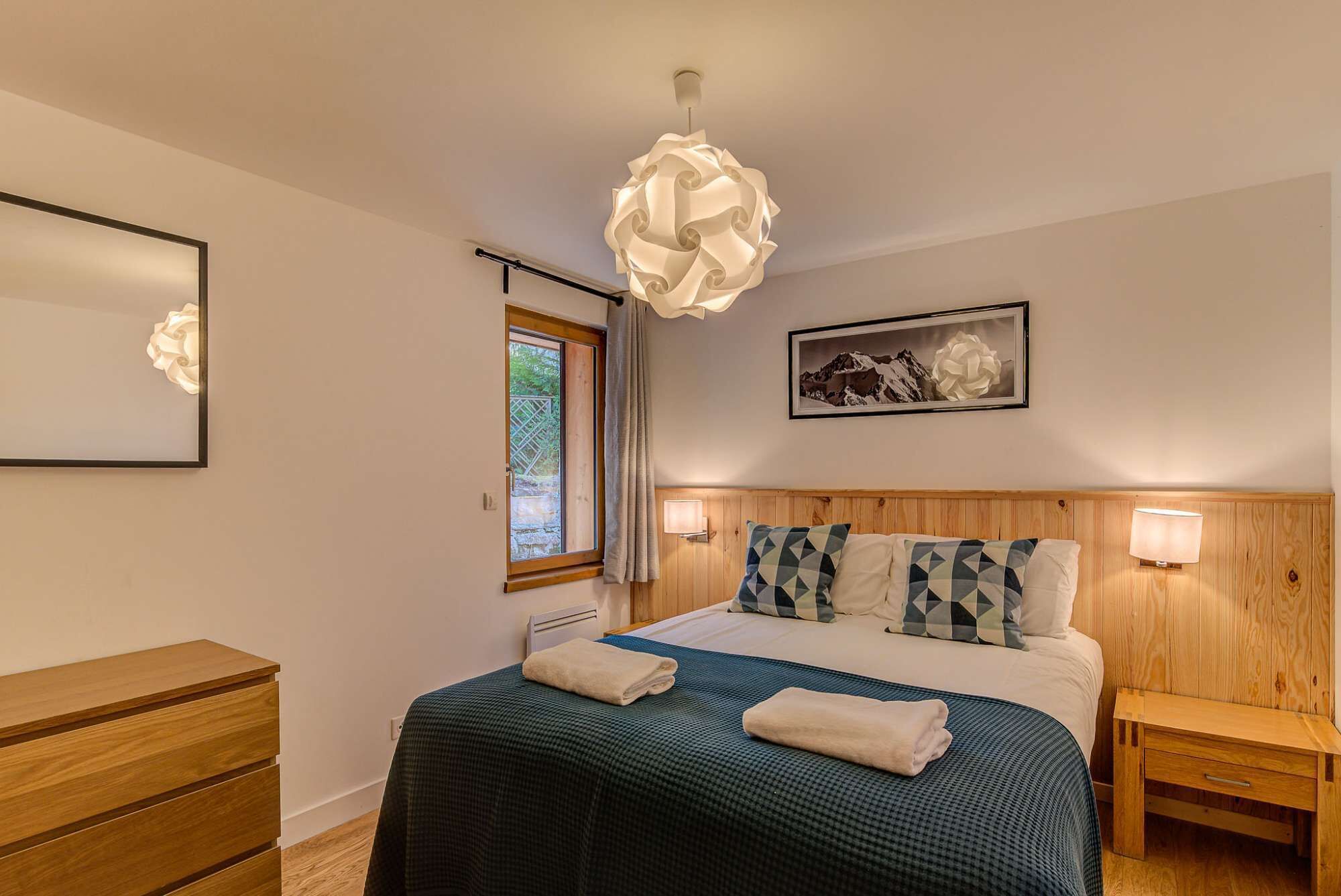 Een slaapkamer met een bed, een houten hoofdbord, nachtkastjes, een raam met gordijnen, een wandlamp en een ingelijst schilderij.