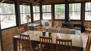 Een houten chalet met een grote eettafel, stoelen, een open haard en grote ramen met uitzicht op een bos.